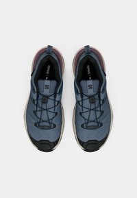 O pereche de pantofi sport albaștri și negri cu tălpi bej și șireturi cu sistem de strângere rapidă, văzuți de sus pe un fundal alb simplu.