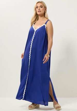 Kvinde iført en ærmeløs koboltblå maxikjole med hvid bølget kant, sideslids og beige platåsandaler, stående mod en ensfarvet baggrund.