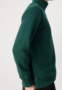 Tmavě zelená mikina se zipem a dlouhými rukávy, ribbenými manžetami a hladkým povrchem. Obsahuje ramenní švy a přiléhavý design.