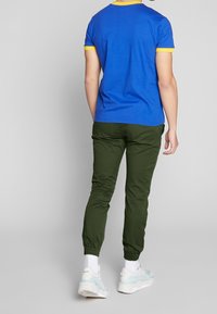 Chemise bleue à manches courtes avec une bordure jaune, associée à un pantalon vert effilé et des chaussures de sport blanches avec des accents bleu clair.