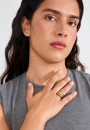 Tume juustega naine, kes kannab varrukateta halli topi, puudutab rinda ja näitab sõrmel laia kuldset rõngast pärliga.