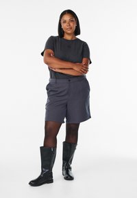Kvinde med glat sort hår står med armene over kors, iført en sort t-shirt, grå shorts, sorte tights og knæhøje sorte støvler.