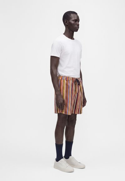 Homme debout portant un t-shirt blanc, un short rayé multicolore, des chaussettes bleu marine et des baskets blanches sur un fond blanc uni.