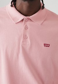 Polo en coton rose avec un col à revers, trois boutons et un petit logo rouge Levi's sur la poitrine gauche. Texture lisse, design décontracté.