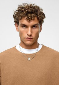 Bruine sweatshirt met een gestructureerd oppervlak, gelaagd over een wit T-shirt. Zilveren ketting met een rechthoekige hanger. Krullend haar.
