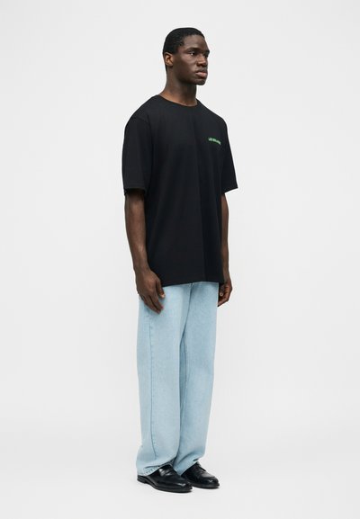 Άνδρας που στέκεται φορώντας μαύρο φαρδύ T-shirt με μικρό πράσινο κείμενο, ανοιχτό μπλε τζιν παντελόνι με φαρδιές σκάλες και μαύρα παπούτσια, μπροστά σε απλό λευκό φόντο.