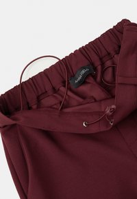 Maroon elastīgas jostas bikses ar saiti un melnu etiķeti iekšpusē, uz balta fona ir rakstīts "ANNA FIELD".