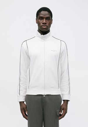 Man wearing a white zip-up jacket with black piping and gray pants with a pocket on the side, standing against a plain background.
