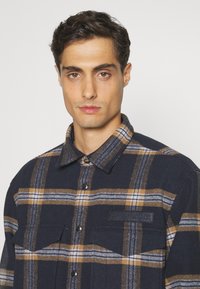 Camicia di flanella blu navy con motivo a quadri giallo e bianco, con bottoni anteriori, colletto a punta e due tasche sul petto. Logo del marchio sulla tasca.