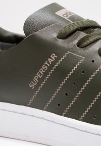 Olivově zelené kožené tenisky s bílou texturovanou gumovou podrážkou, akcentovými pruhy ve tvaru cik-cak a perforacemi pro lepší prodyšnost.