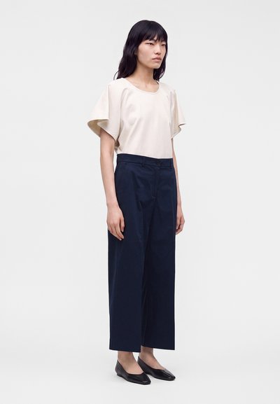 Femme portant un chemisier crème à manches courtes, un pantalon large bleu marine et des ballerines noires, debout devant un fond clair uni.