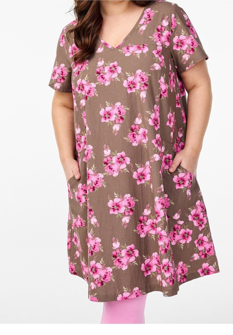 Femme portant une robe marron à col en V avec un imprimé floral rose, manches courtes et poches, mains dans les poches, associée à des leggings roses.
