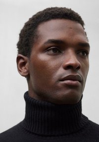 Schwarzer gerippter Rollkragenpullover aus weichem Material, mit hohem Kragen und enganliegendem Schnitt, getragen von einem Modell mit kurzen Haaren.