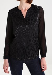Blouse noire à motif léopard, dotée de manches longues transparentes, d'un décolleté en V et d'un tissu texturé. Portée avec un jean foncé.