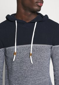 Marineblauwe en grijze hoodie met een gebreide textuur, witte trekkoorden en gouden accenten nabij de opening van de capuchon.