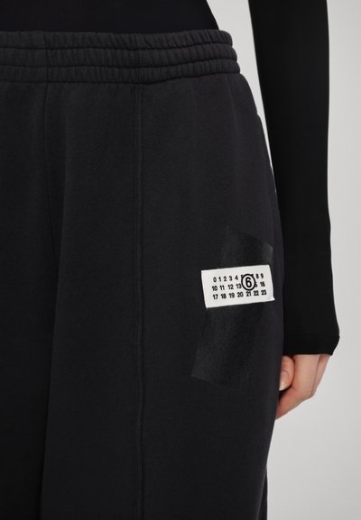 Svarta sweatpants med elastiskt midjeband, sidoficka med en vit etikett som visar siffror, och en strukturerad tygdetalj.