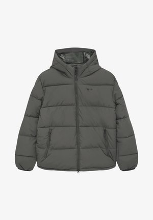 Persona che indossa una giacca imbottita grigio scuro sopra una camicia a quadri rossa e bianca e una maglia nera, abbinata a pantaloni bianco sporco, con le mani in tasca.