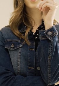 Giubbotto di jeans in tessuto blu scuro, con quattro tasche frontali, dettagli in bottoni e cuciture visibili. Indossato sopra una camicia nera con motivo floreale.