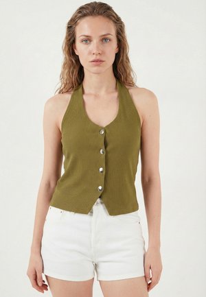 Kvinde iført olivengrøn ærmeløs haltertop med sølvknapper og hvide højtaljede shorts, stående foran en ensfarvet baggrund.