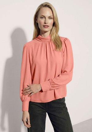 Vrouw met lang blond haar die een koraalroze blouse met lange mouwen en een zwarte broek draagt, staand tegen een bleke achtergrond.