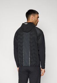Veste de sport noire avec capuche, présentant des motifs géométriques texturés et des accents réfléchissants à l'arrière. Fabriquée en matériau léger.