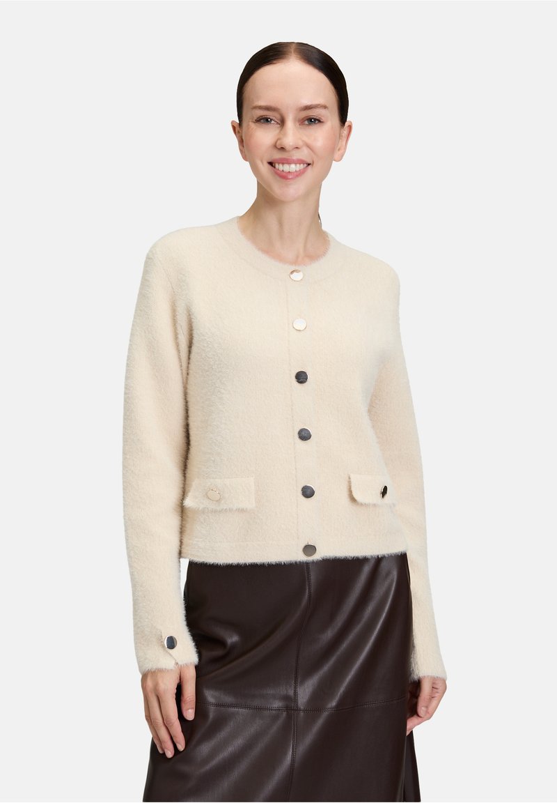 Kobieta uśmiechająca się, ubrana w kremowy sweter na guziki z przednimi kieszeniami i ciemnobrązową skórzaną spódnicę, stojąca na jednolitym tle.