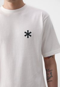 T-shirt en coton blanc à manches courtes, avec un logo astérisque noir sur la poitrine. Texture lisse, coupe décontractée.