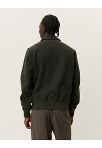 Giacca bomber verde oliva con colletto, polsini a costine e orlo a costine. Tessuto morbido, texture liscia e dettagli minimi. Indossata sopra pantaloni a fantasia.