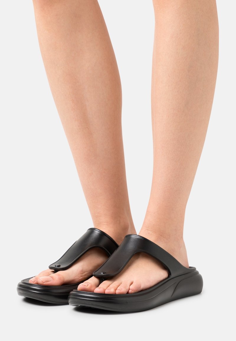 Pieds portant des sandales à lanières noires rembourrées avec des semelles épaisses sur une surface gris clair.