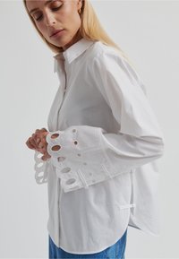 Chemise blanche boutonnée avec manches longues présentant des découpes circulaires aux poignets, en tissu léger, associée à un jean en denim bleu.