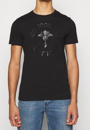 Mann trägt schwarzes T-Shirt mit kurzen Ärmeln mit rundem geprägtem Logo und Symbol, kombiniert mit blauen Jeans vor hellem Hintergrund.