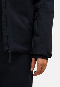 Marineblaue wasserdichte Jacke mit Reißverschluss, verstellbaren Bündchen mit Klettverschluss und glatter matter Textur. Präsentation eines eng anliegenden Ärmels.