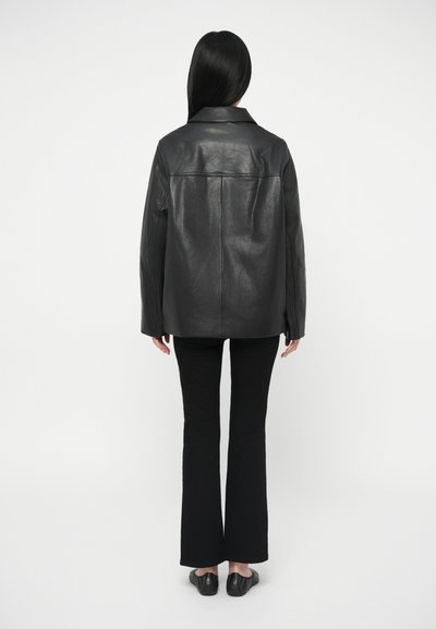 Άτομο με μακριά μαύρα μαλλιά, φορώντας μαύρο δερμάτινο μπουφάν, μαύρο φαρδύ παντελόνι και μαύρα παπούτσια χωρίς λουριά, φωτογραφημένο από πίσω σε λευκό φόντο.