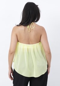 Mujer con cabello largo y oscuro que lleva una blusa sin mangas, suelta, color amarillo claro y pantalones negros, de espaldas contra un fondo liso.