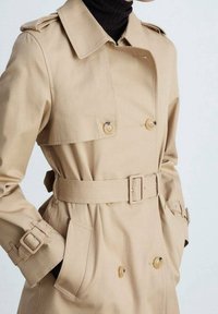 Manteau trench beige avec un design à double boutonnage, grands boutons, taille ceinturée et poignets ajustables. Tissu lisse avec un léger éclat.