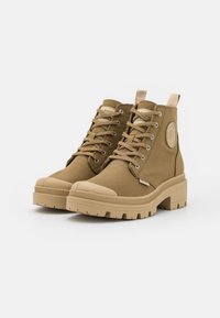 Botas de tobillo de lona color arena con punteras de goma y suelas texturizadas. Cuentan con cordones, una pestaña trasera y un detalle de logotipo circular en el lateral.