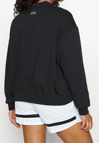 Sudadera negra de gran tamaño con cuello redondo y logo bordado en la parte posterior, combinada con pantalones cortos blancos con rayas negras.