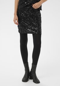 Fekete flitteres mini szoknya, amely testhezálló kialakítással és texturált felülettel rendelkezik, fekete átlátszatlan leggingsszel és bokacsizmával párosítva.
