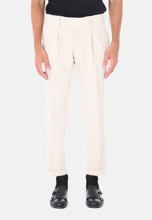 Pantaloni beige a plissettatura con vestibilità su misura, realizzati in un tessuto liscio e con orlo risvoltato, abbinati a mocassini neri e calze.