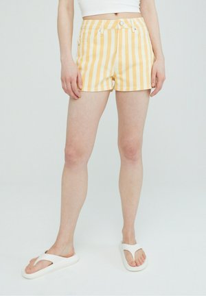 Persoon die een korte broek met gele en witte verticale strepen draagt, een wit crop top en witte slippers, staande tegen een effen lichte achtergrond.