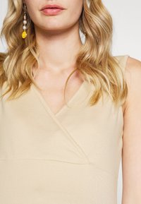 Top beige com decote em V, feito de um tecido suave. Características notáveis incluem alças finas e um silhueta ajustada.
