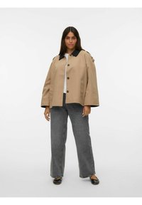 Beige regnjakke med sort krave og knapper, med en løs pasform og sidelommer, parret med grå vide bukser og sorte flade sko.