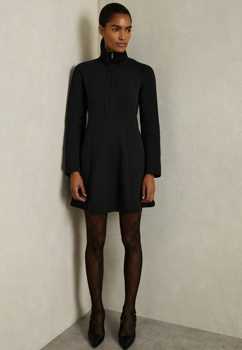 Schwarzes Kleid mit hohem Kragen und Reißverschluss, das einen taillierten Oberteil und einen ausgestellten Rock hat. Tragen mit schwarzen, diamantgemusterten Strumpfhosen und spitzen Absätzen.