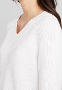 Pull blanc en maille avec un col en V et des détails côtelés. La texture semble douce et légère, adaptée aux tenues superposées.