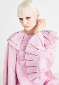 Ροζ φόρεμα με μακριά μανίκια που διαθέτει λαιμόκοψη και μανίκια με ρυτίδες, φτιαγμένο από ένα απαλό ύφασμα με ματ φινίρισμα. Ασυμμετρία στη θέση των ρυτίδων.