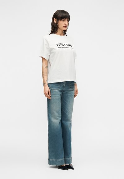 Άσπρο κοντομάνικο μπλουζάκι με μαύρο κείμενο «ΕΙΝΑΙ ΕΝΤΑΞΕΙ Βάλε απλώς μια λάμψη επάνω του», συνδυασμένο με φαρδιά ανοιχτό μπλε τζιν και μαύρα μυτερά παπούτσια.