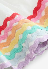 Filas de cintas onduladas coloridas y texturizadas cosidas sobre tela blanca, dispuestas en diagonal en tonos rojo, rosa, naranja, amarillo, verde, azul y púrpura.