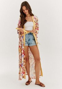 Pestrobarevný květinový kimono s žlutými lemy a bočními šňůrkami, nošené přes bílé krátké tričko a džínové šortky, vyrobené z lehkého a vzdušného materiálu.