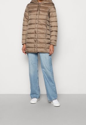 Personne portant une veste matelassée marron clair, un jean large bleu clair et des baskets blanches, debout devant un fond blanc uni.