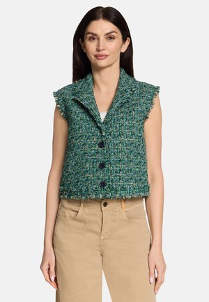 Femme portant un gilet texturé sans manches vert et bleu avec des boutons et des bords effilochés, un pantalon beige et un top blanc en dessous.